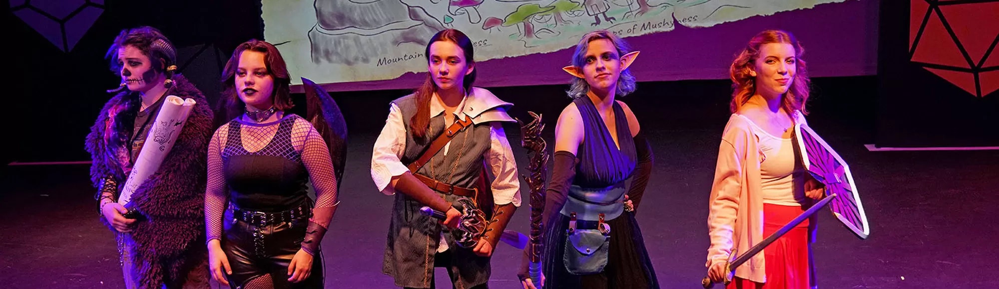 Five people in fantasy costumes and makeup on stage, with a colorful backdrop map.