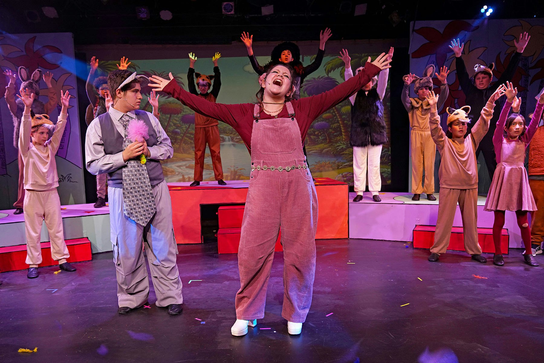 A lively stage performance featuring actors in playful costumes, with one prominently singing and several others participating.