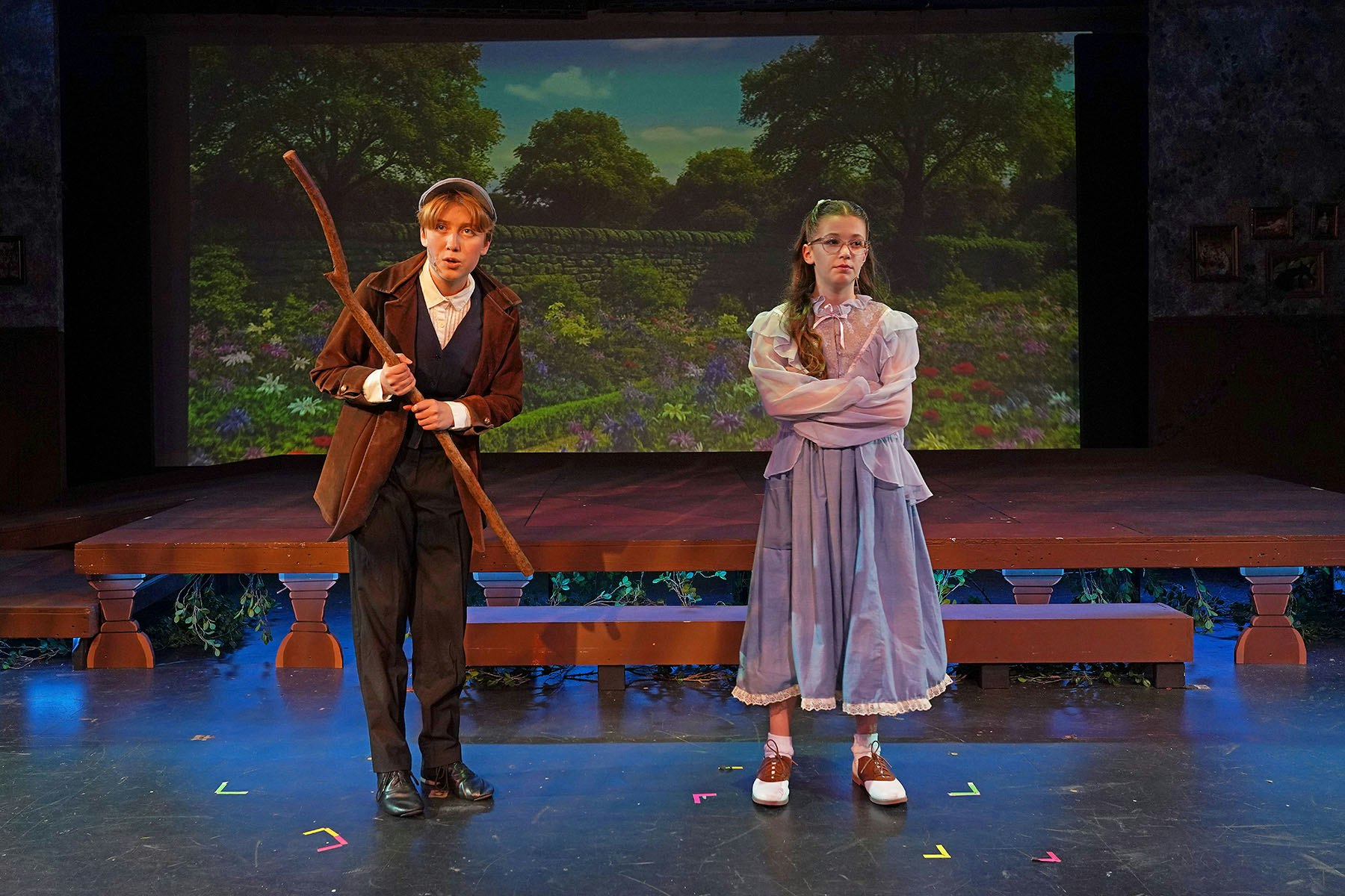 Two people in period clothing perform onstage with a forest backdrop; one holds a stick.