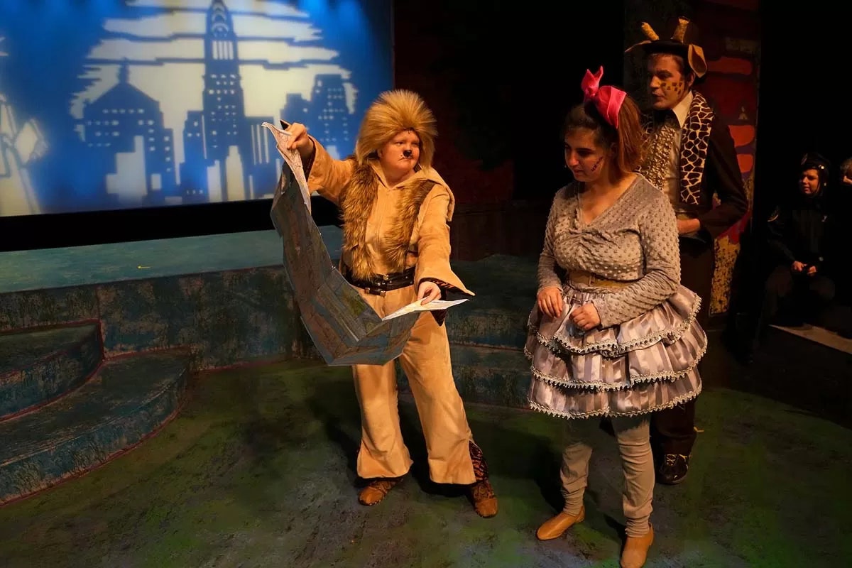 Three people in animal costumes on stage; one is holding a map.