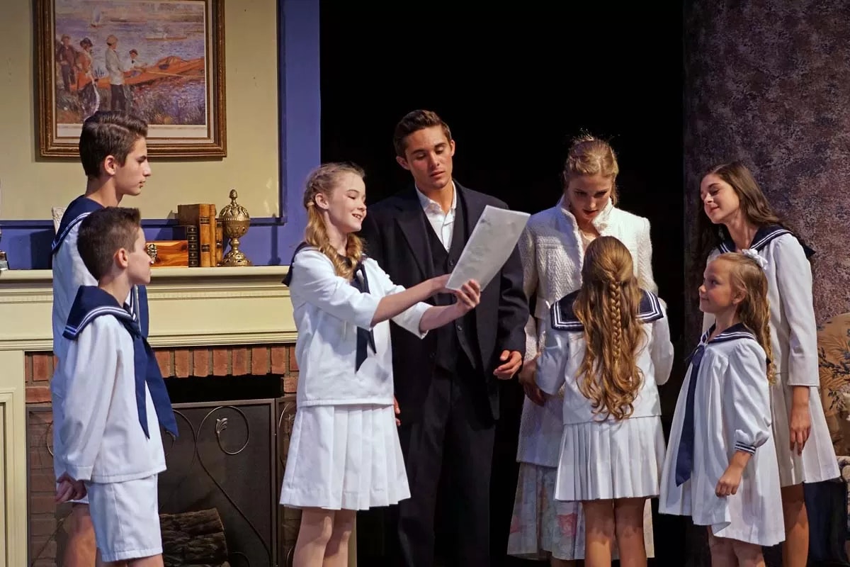 A group of people, some in sailor uniforms, stand together on stage. One holds a paper, others gather around, suggesting a theatrical scene.
