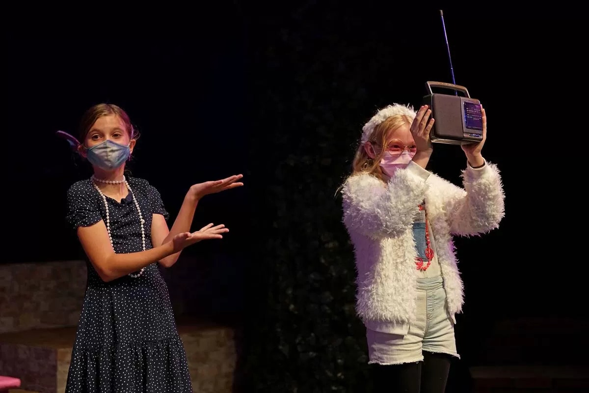 Two children on stage, wearing masks; one holds a radio, the other gestures with open hands.