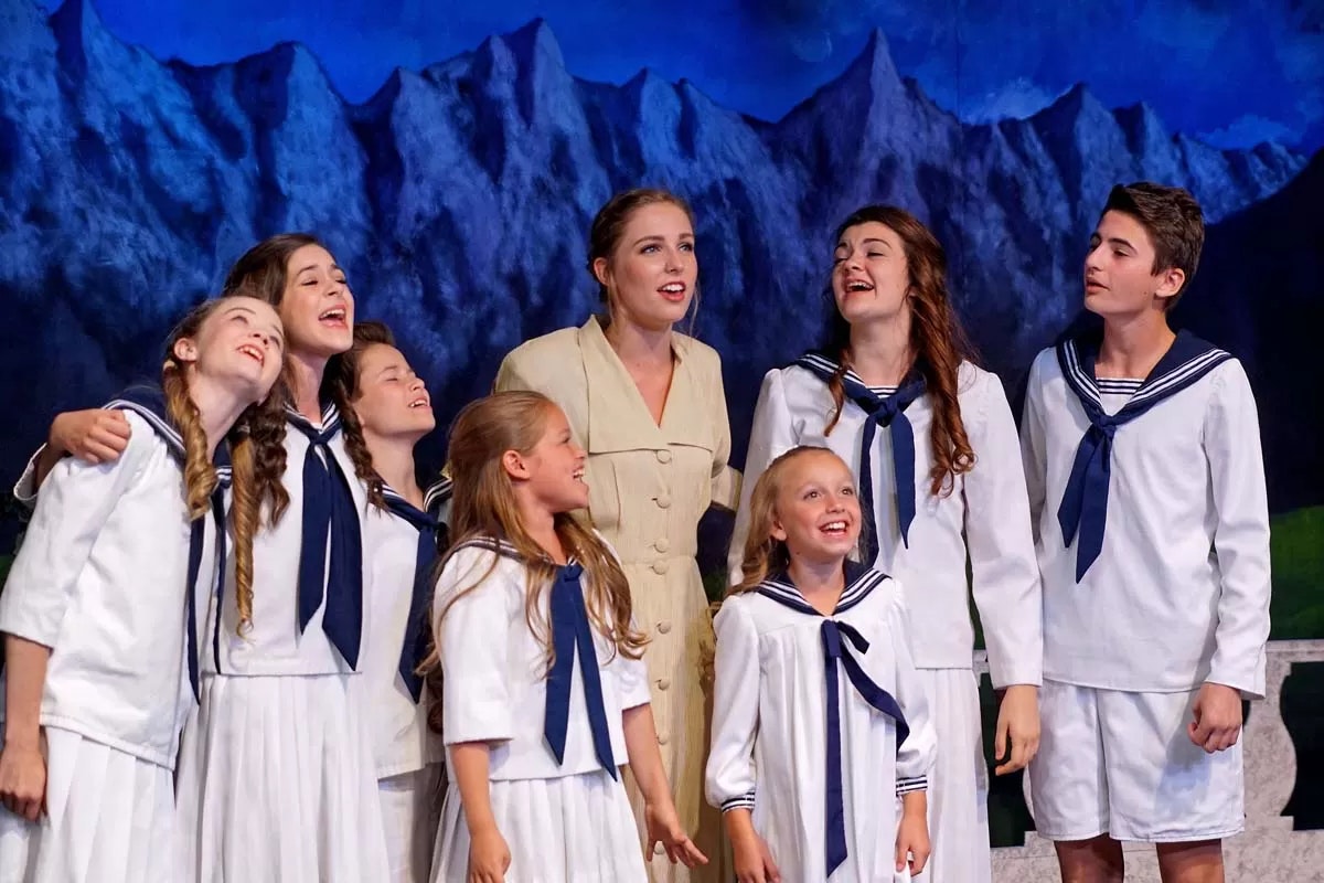 Seven children in sailor outfits and an adult on stage, with mountains painted in the background, all appear to be singing.