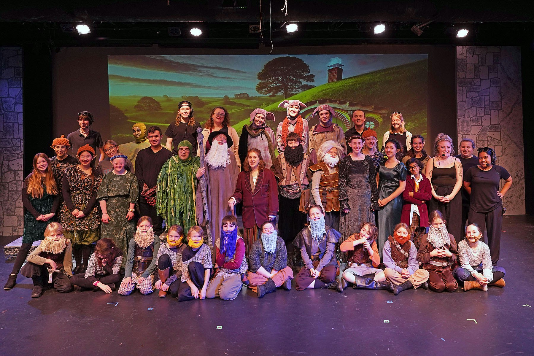 A group of people in costumes posing on stage, resembling a cast from a theater production.