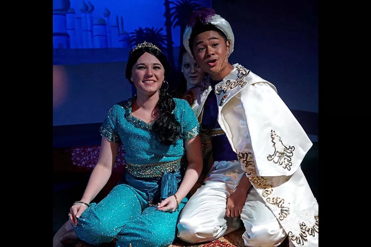 Two people in colorful costumes on stage, one in blue-green and the other in white with gold trim, set against a fantasy backdrop.