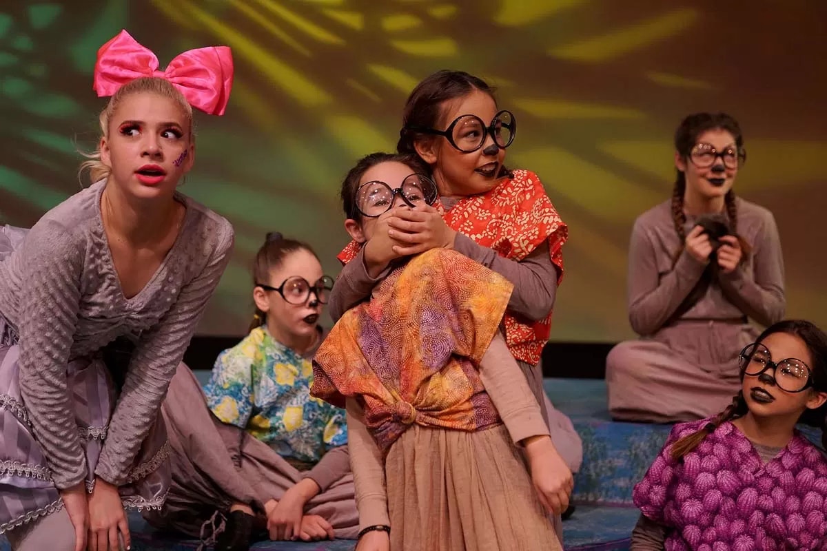Children in colorful costumes and large glasses performing on stage, one with a large pink bow and others with expressive makeup.