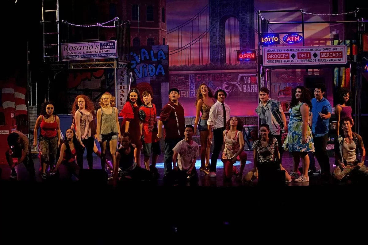 A group of performers on stage with a colorful urban backdrop, dressed in various outfits, suggesting a lively theatrical scene.