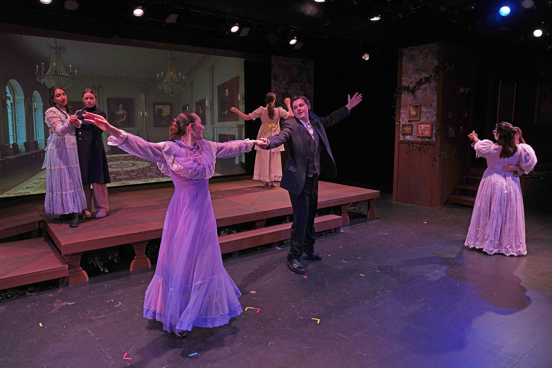 A stage scene with people in period costumes dancing and interacting amidst a dramatic background.