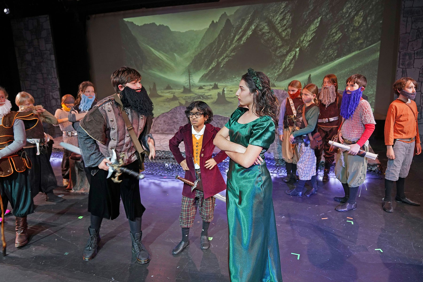 A group of children in costumes perform on stage with a fantasy backdrop, appearing to be in a theatrical play.