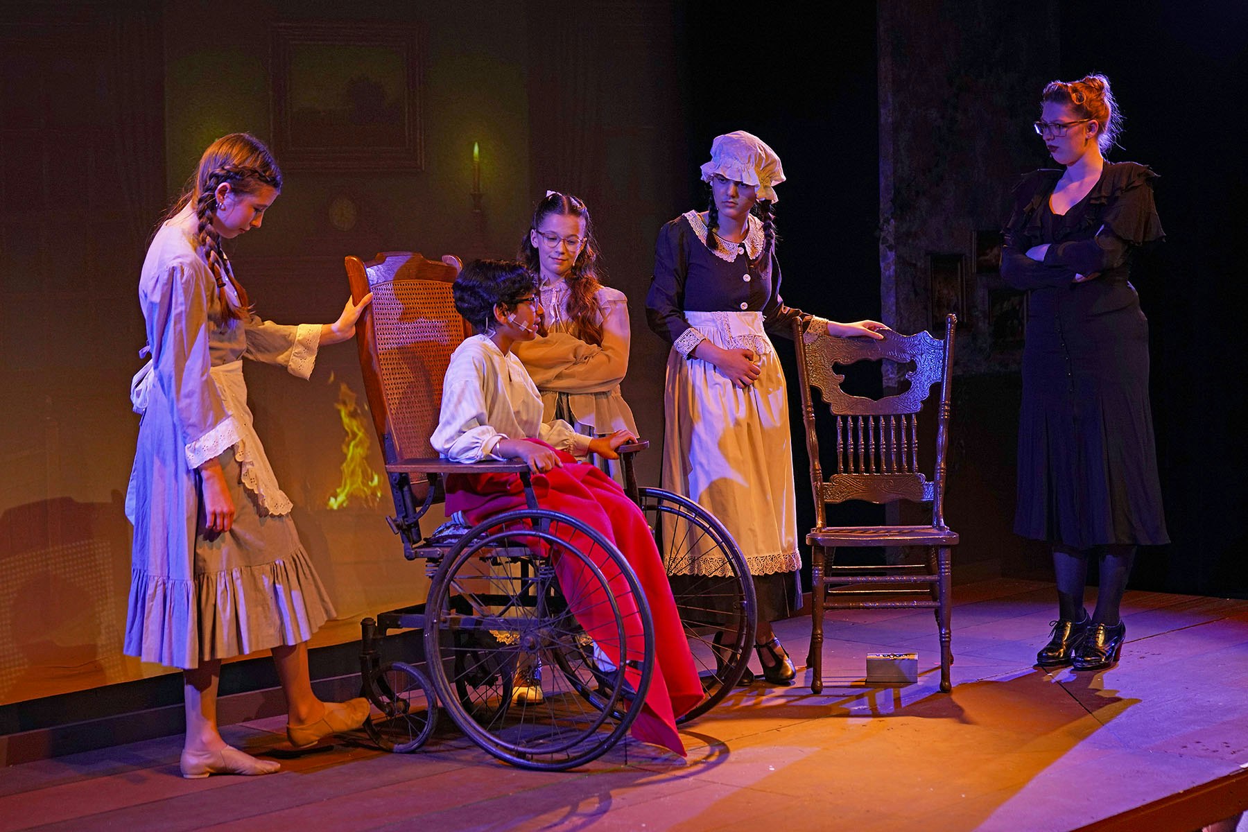 A stage scene with five actors in period costumes; one is in a wheelchair.
