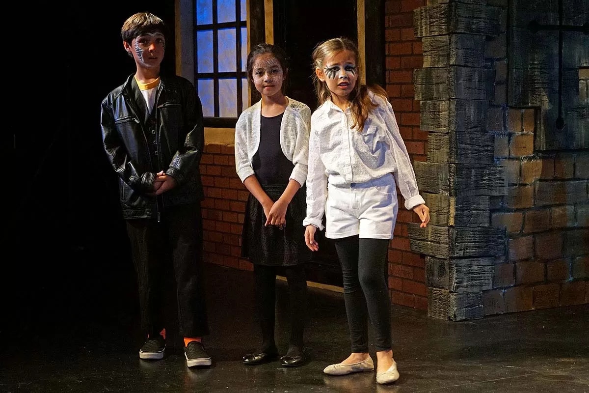 Three children in theatrical costumes and makeup on stage, standing before a brick backdrop, possibly performing a play.