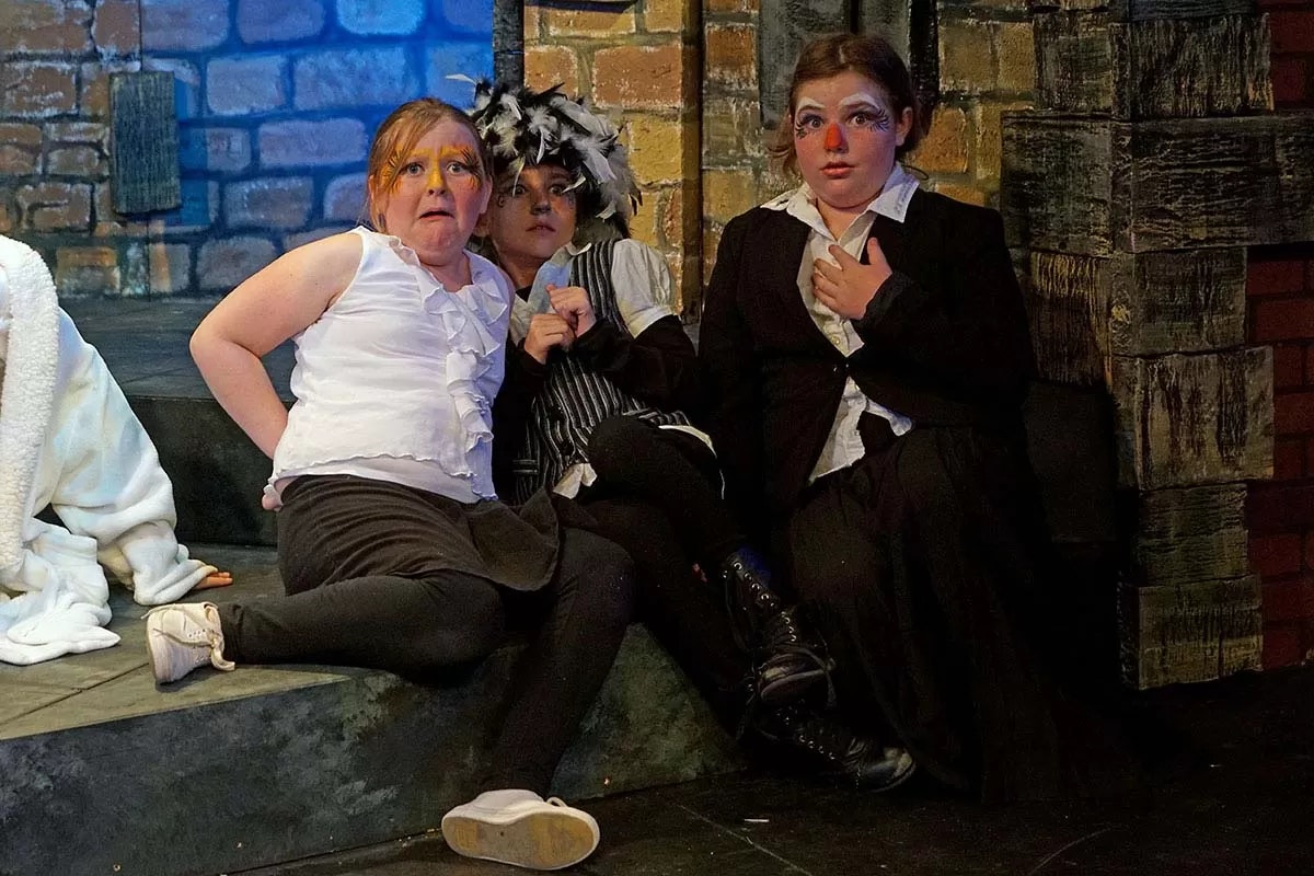 Three children in theatrical costumes and makeup, sitting against a stone wall.