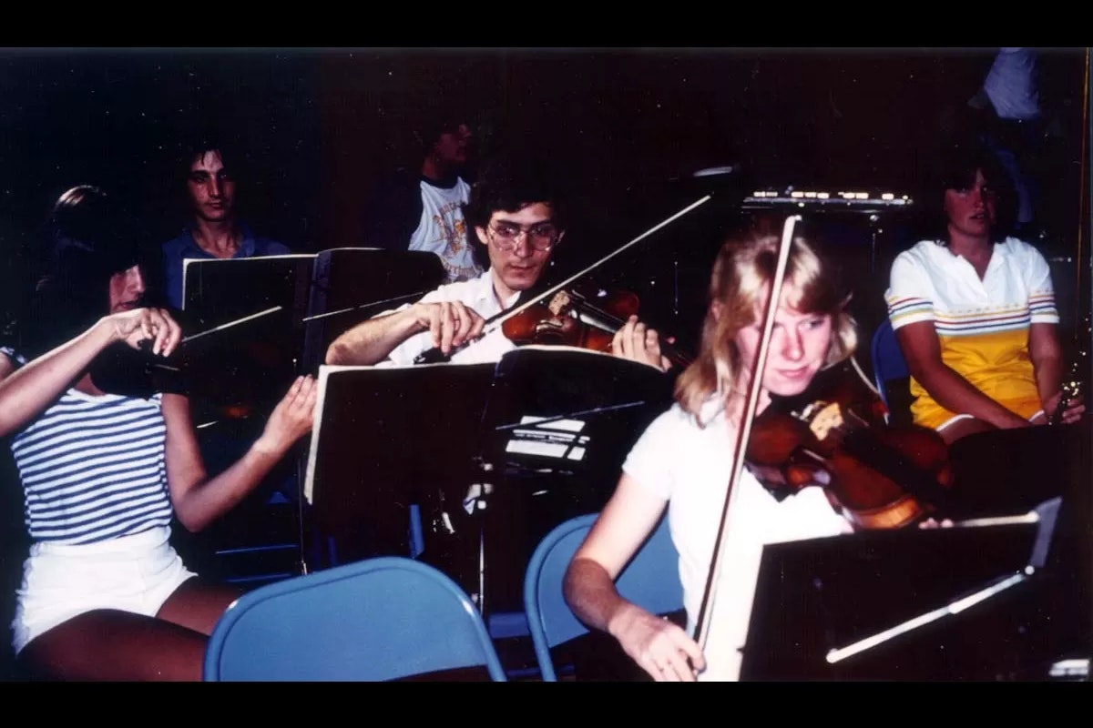 People playing violins in an orchestra setting, seated on blue chairs with music stands in front of them.