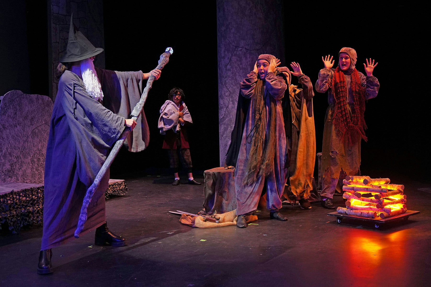 A wizard brandishes a staff at three frightened people near a glowing faux campfire on a stage.