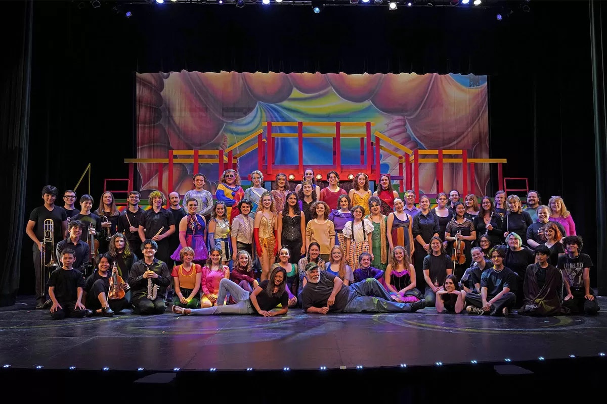 A large group of people on a stage with colorful costumes and a painted backdrop, likely a theater cast and crew.