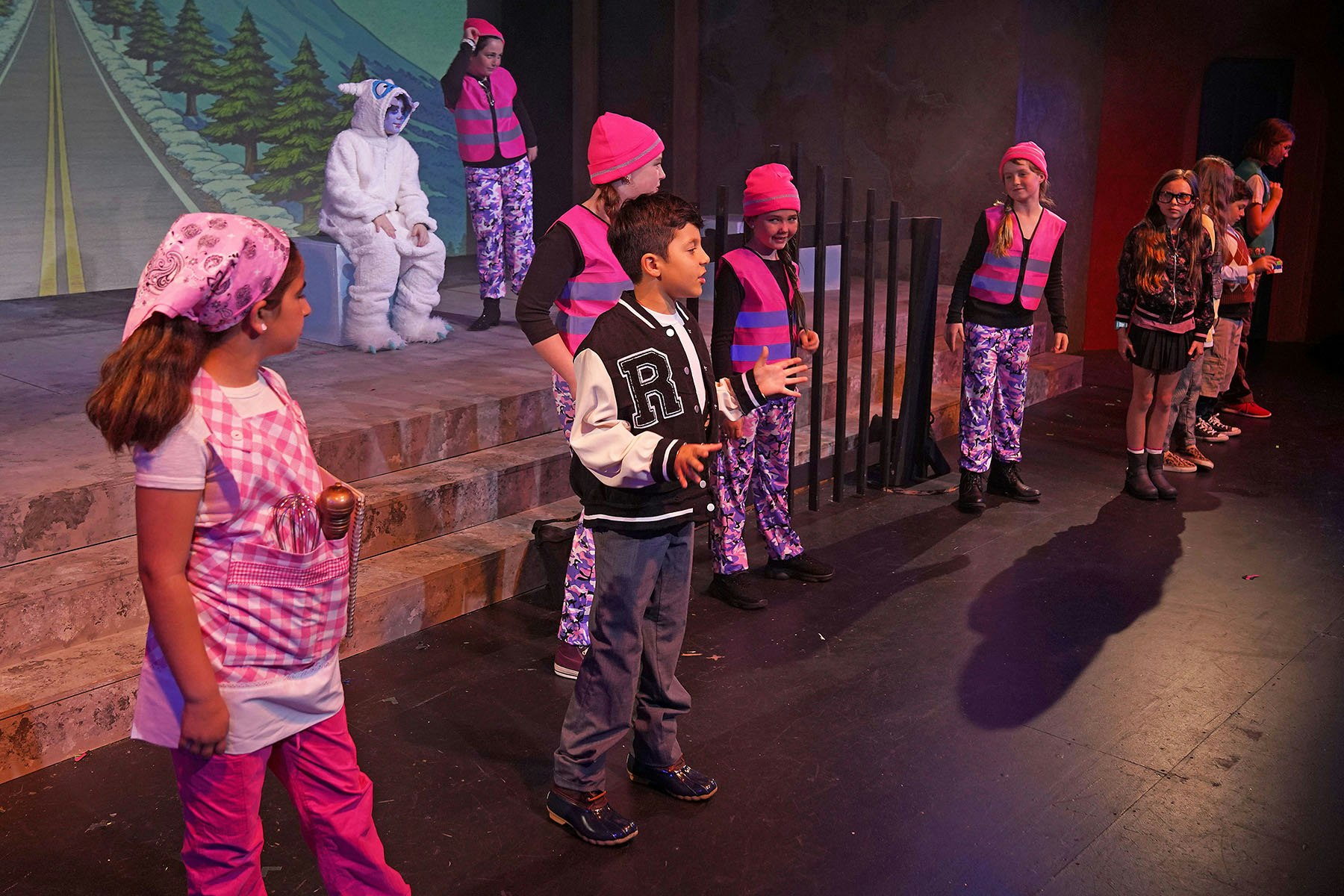 A group of children performing on stage, wearing colorful costumes, with a backdrop depicting a road and trees.