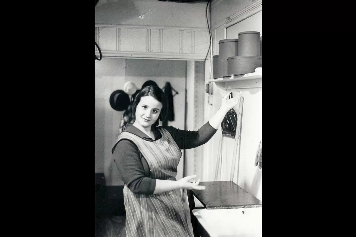 A woman in a striped apron gestures beside a shelf with containers in a kitchen-like setting.