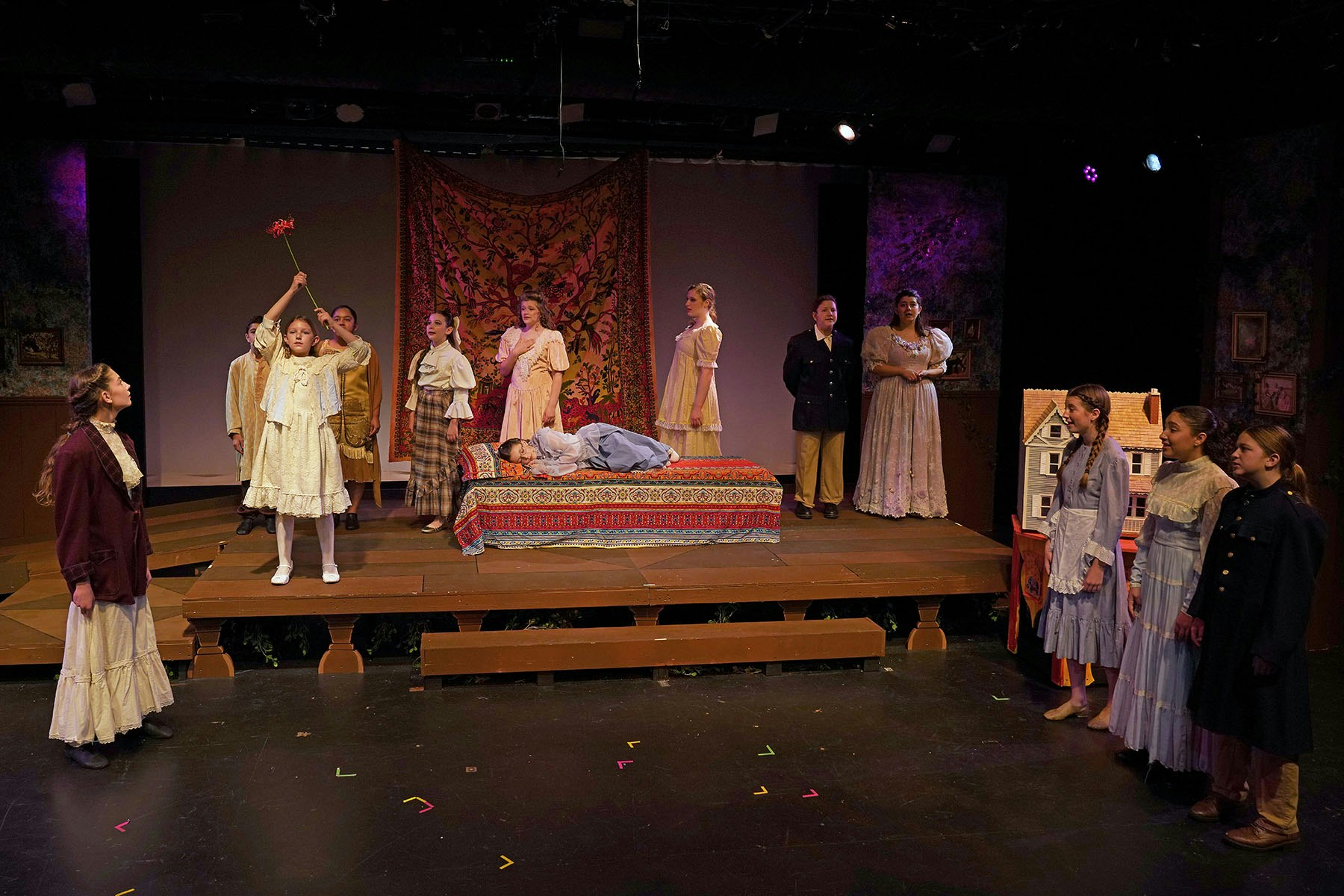 Theatrical scene with actors in period costumes on stage, one holding a wand, another lying on a bed, surrounding cast watching.