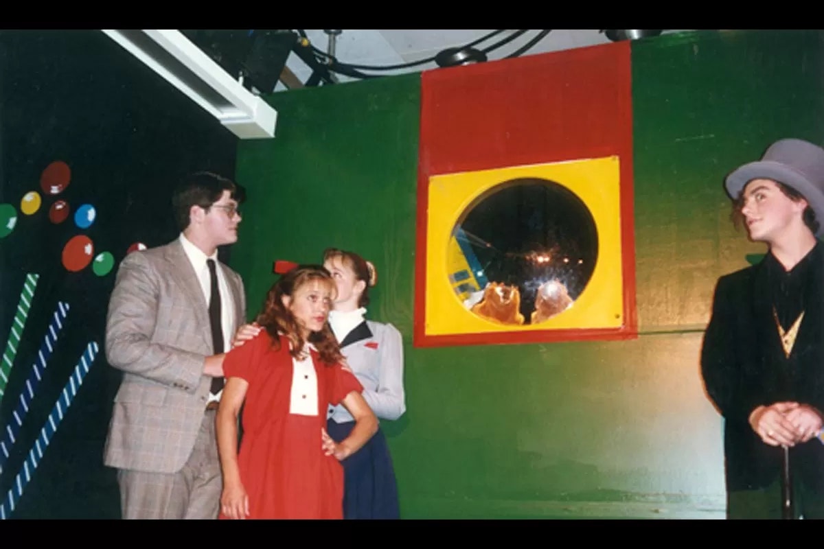 A group of people in a colorful room, with a person dressed in a top hat and suit on the right, and reflections in a round window.