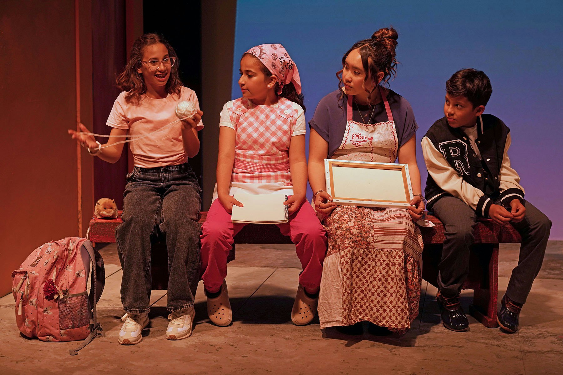 Four children sit on a bench, engaging with each other. One holds a ball and others have notebooks or blank boards, in a vibrant setting.
