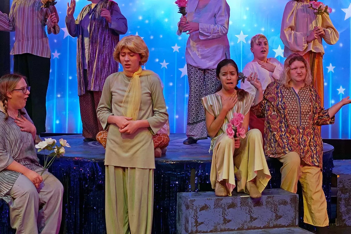 A group of people performing on stage, wearing colorful clothes, holding flowers, against a starry backdrop.