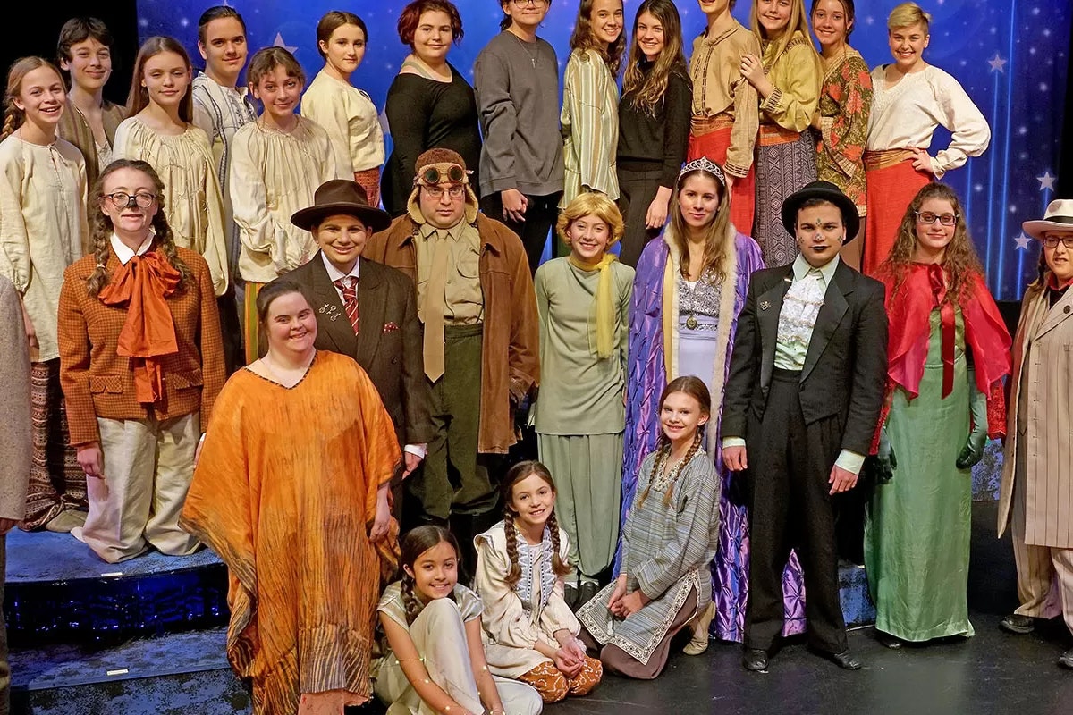 A large group of people in theatrical costumes posing on a stage with a blue starry backdrop.