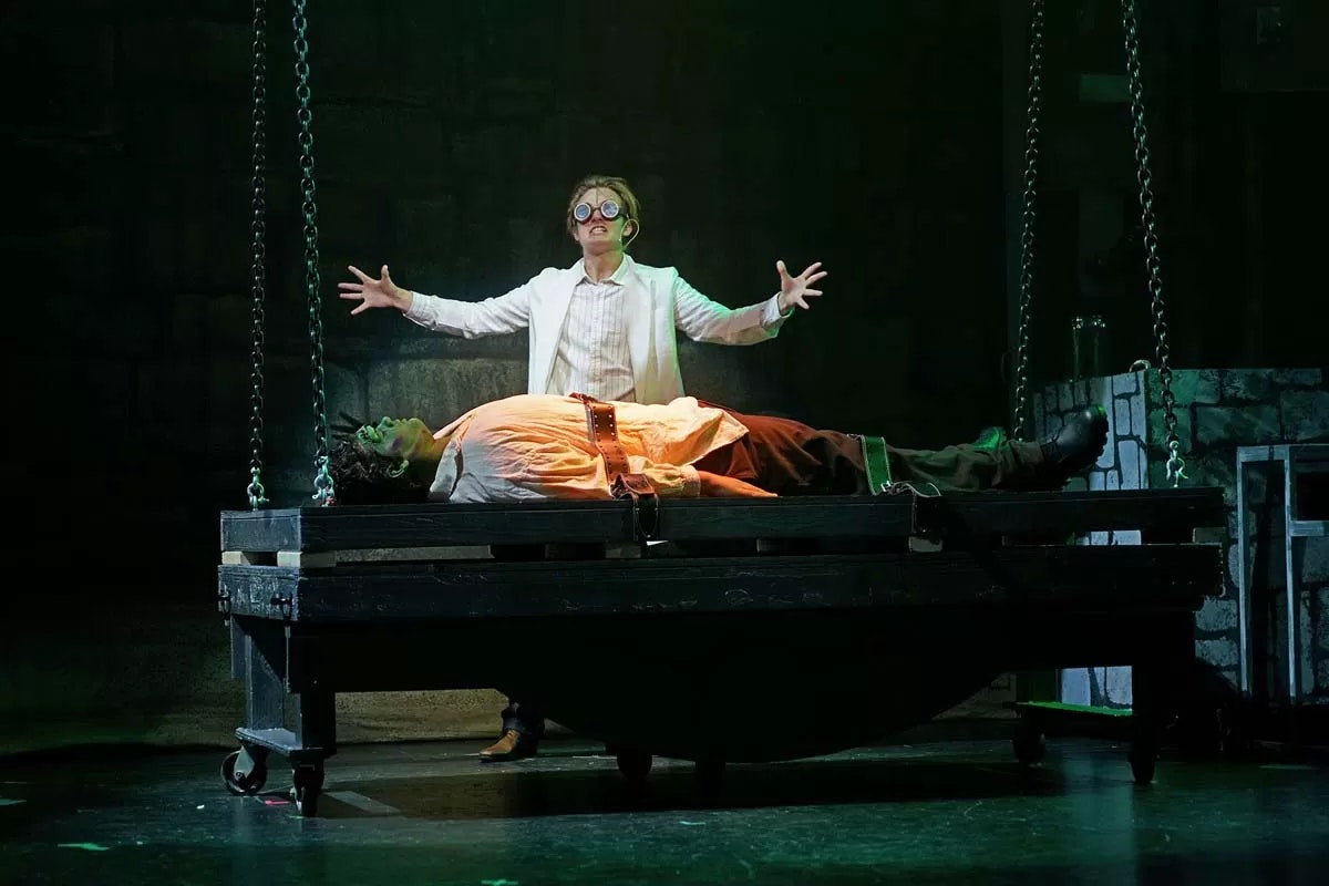 A person in a lab coat and goggles stands over another lying on a table, in a dramatic, theatrical scene.