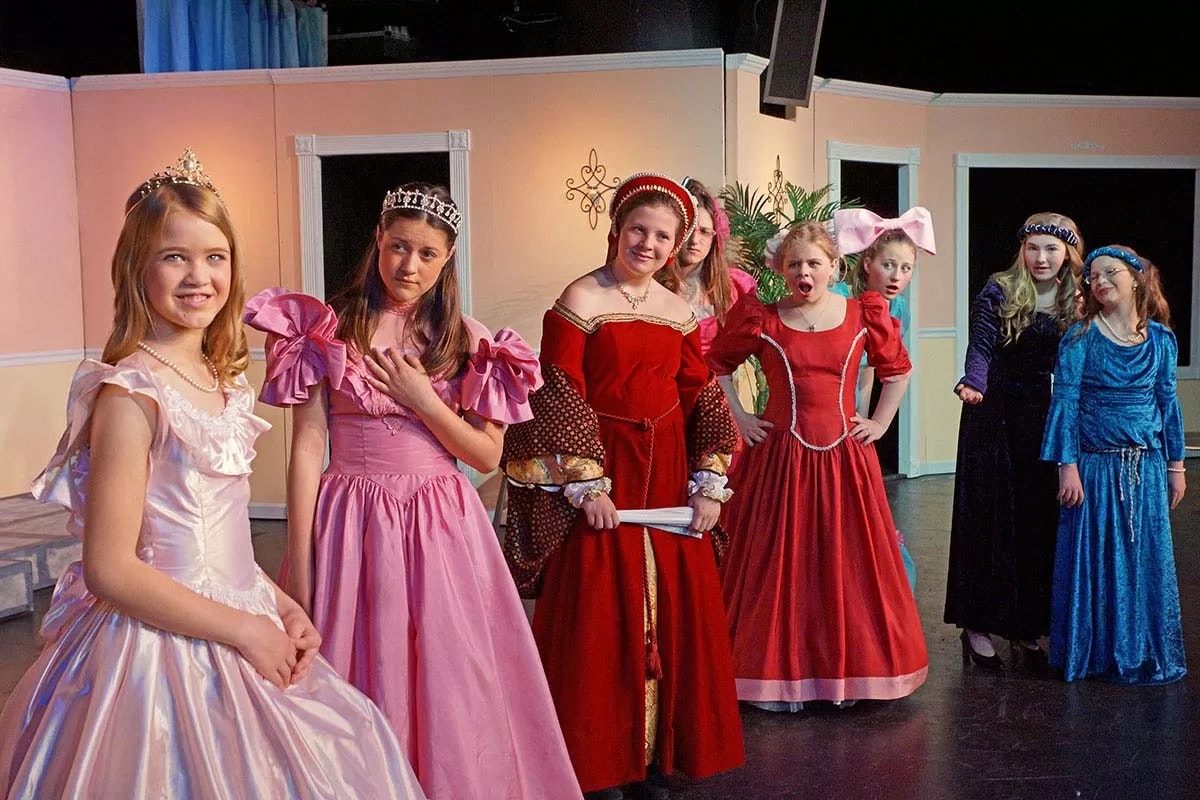 A group of children in costumes on stage, dressed as princesses, standing together and smiling.