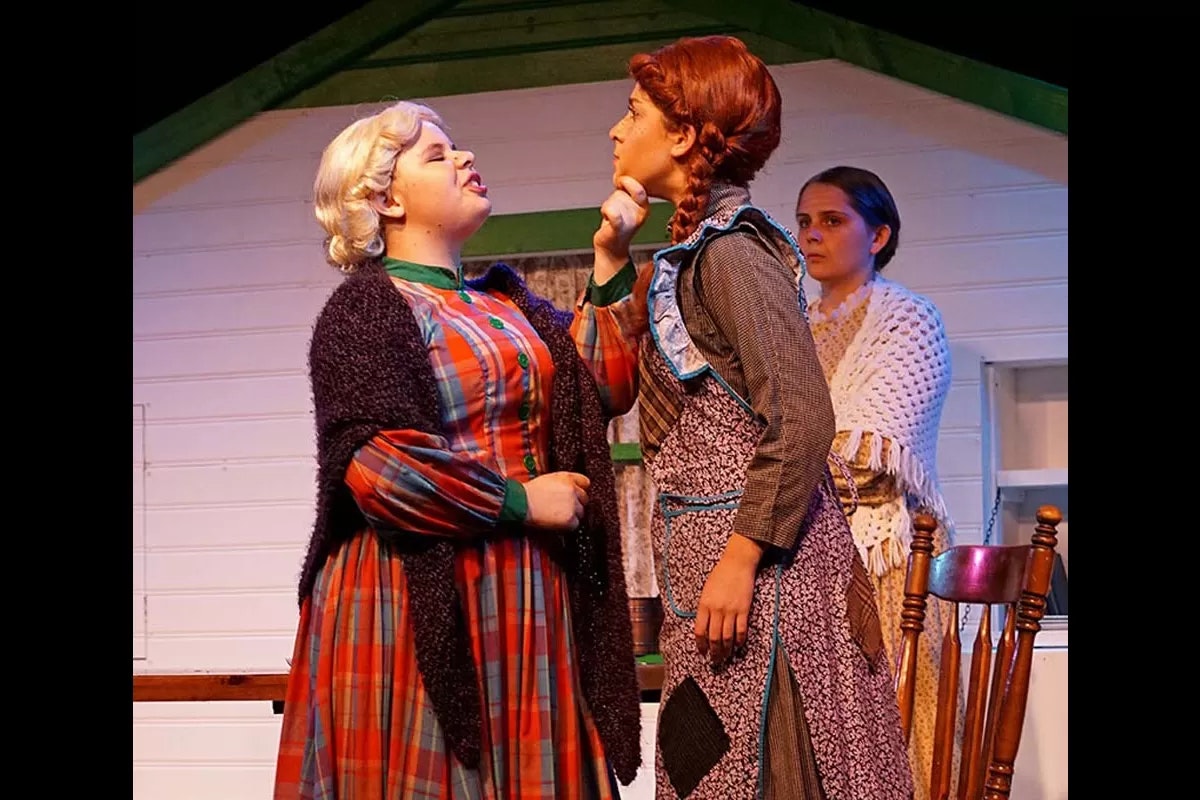Three people in a theatrical scene; one is gesturing towards another, and the third watches in the background.