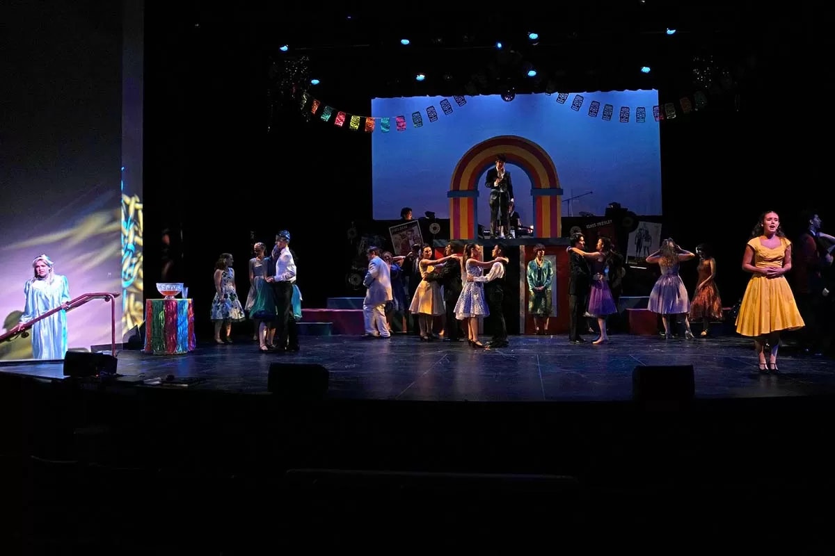 A stage performance with people dancing, a colorful backdrop, and a woman in a yellow dress singing.