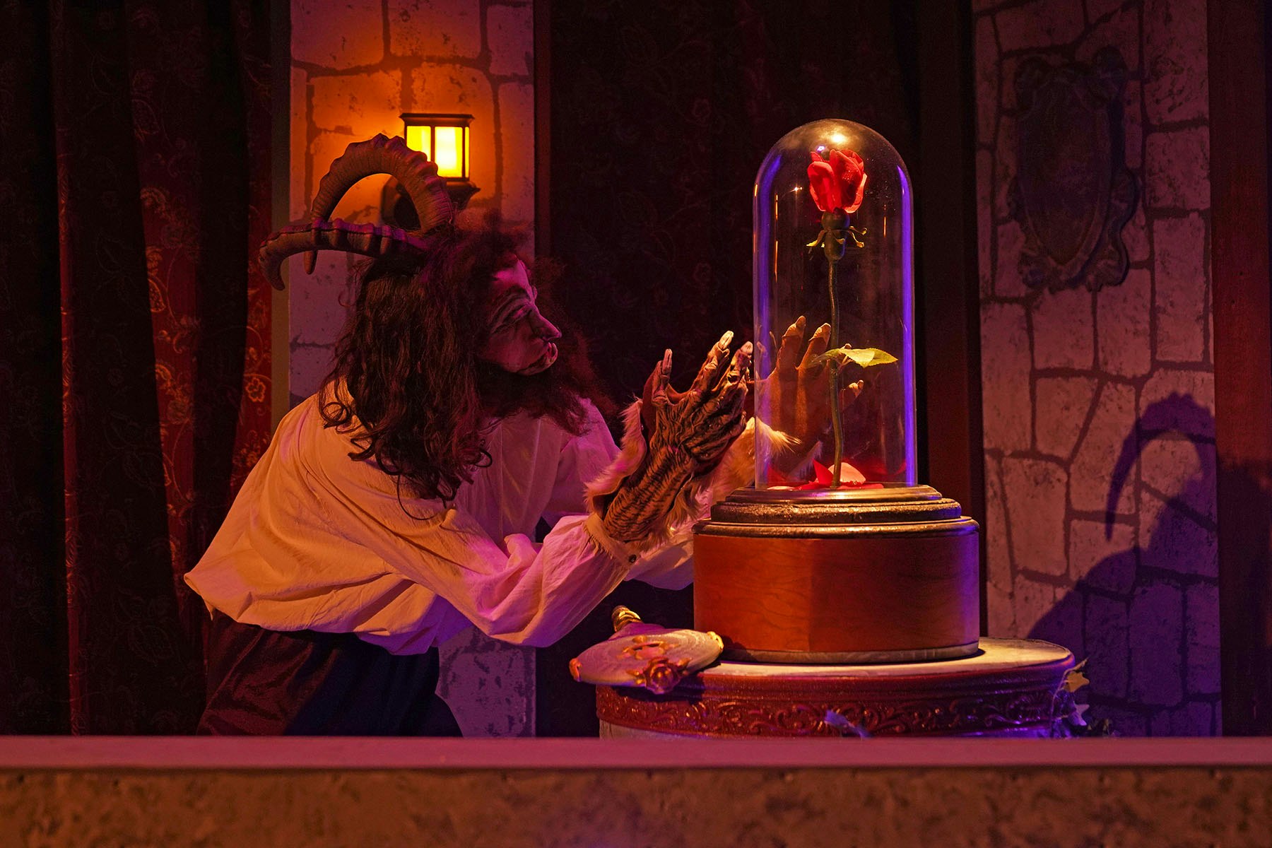 A costumed person resembling a beast interacts with a glowing red rose under a glass dome in a dramatic setting.