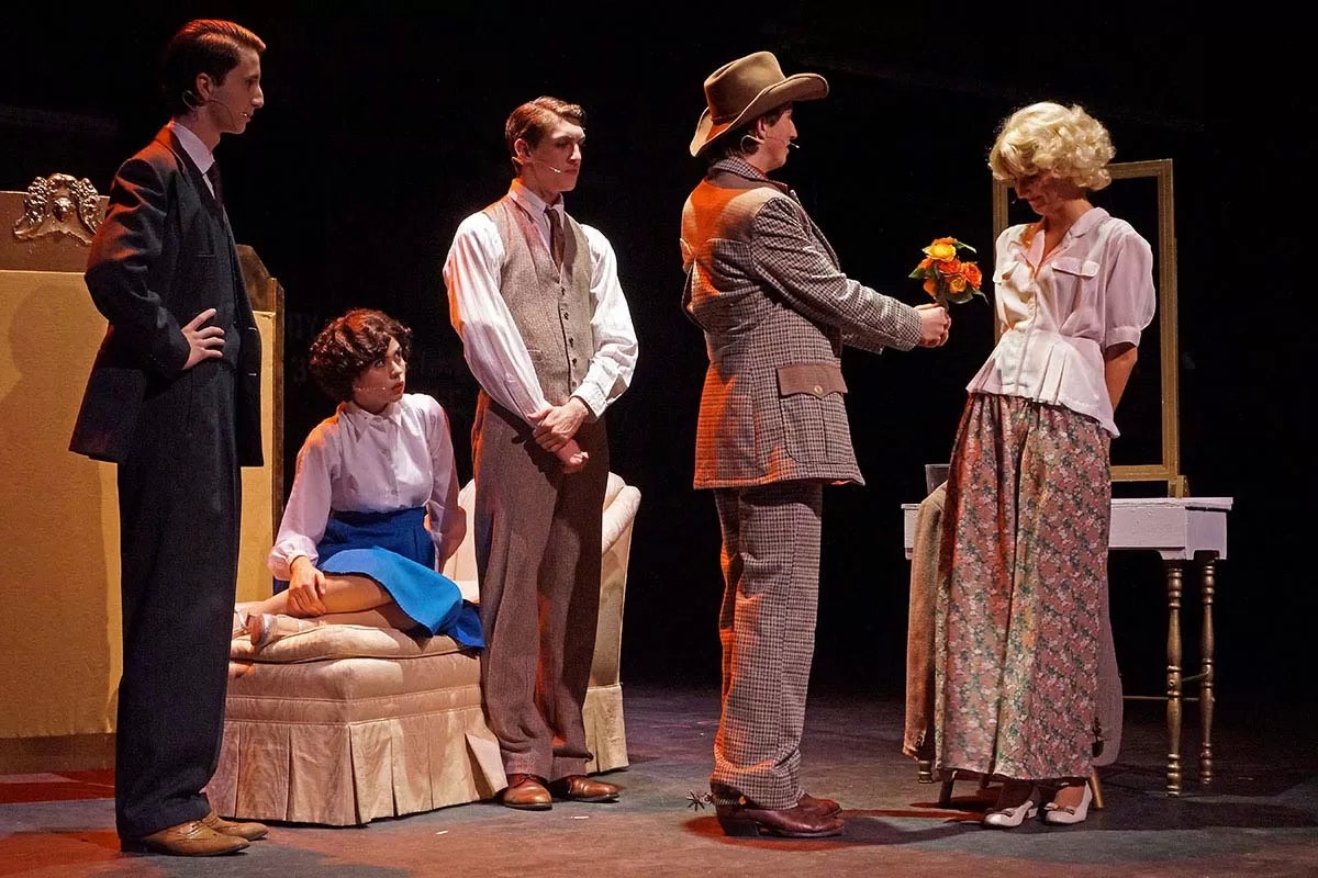 Five people in period costumes on stage, one man offers flowers to a woman, seated woman observes.