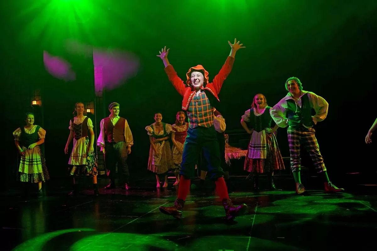 Performers in colorful costumes on stage under green light, with one person in front striking an expressive pose.