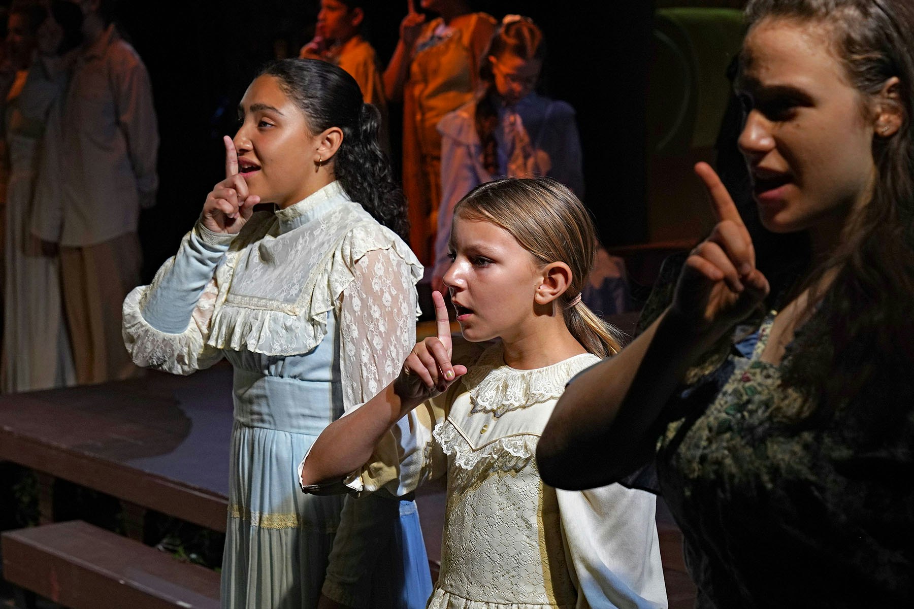 People in period costumes on stage making a "shush" gesture.