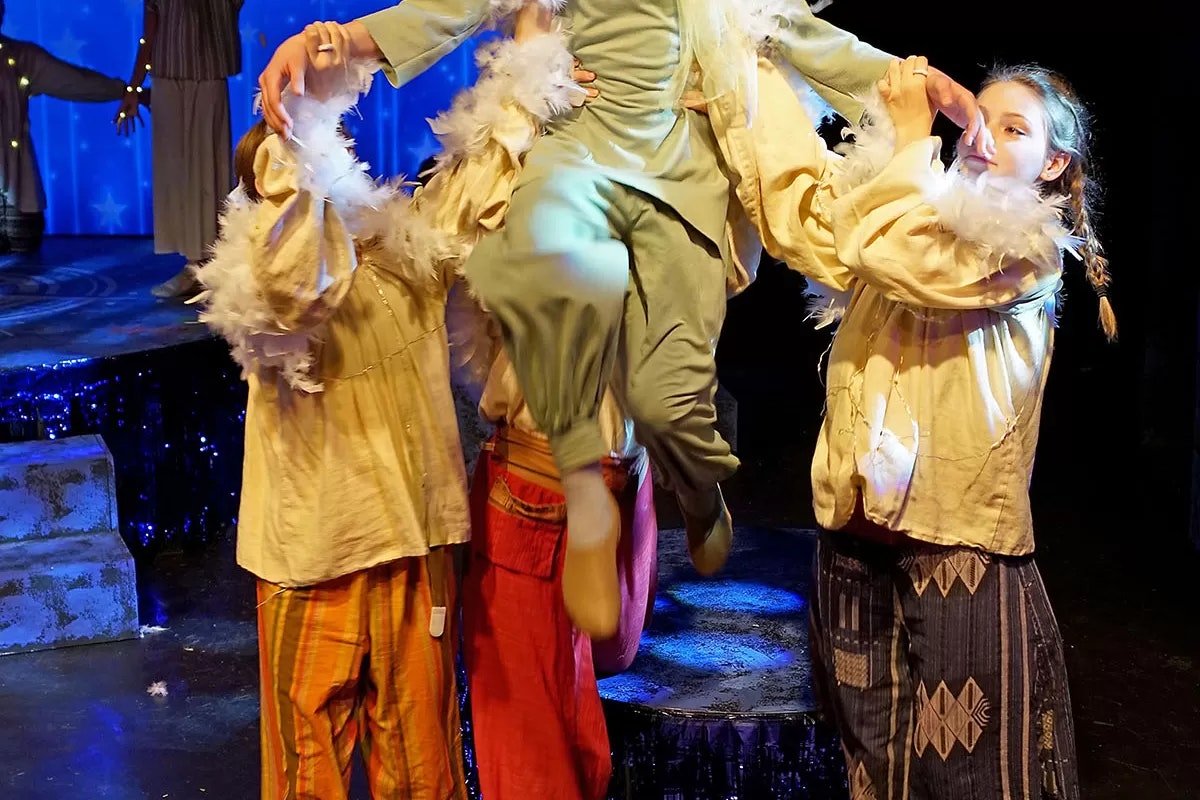 A group of actors on stage, with one person being lifted, wearing colorful costumes and feather details against a blue backdrop.