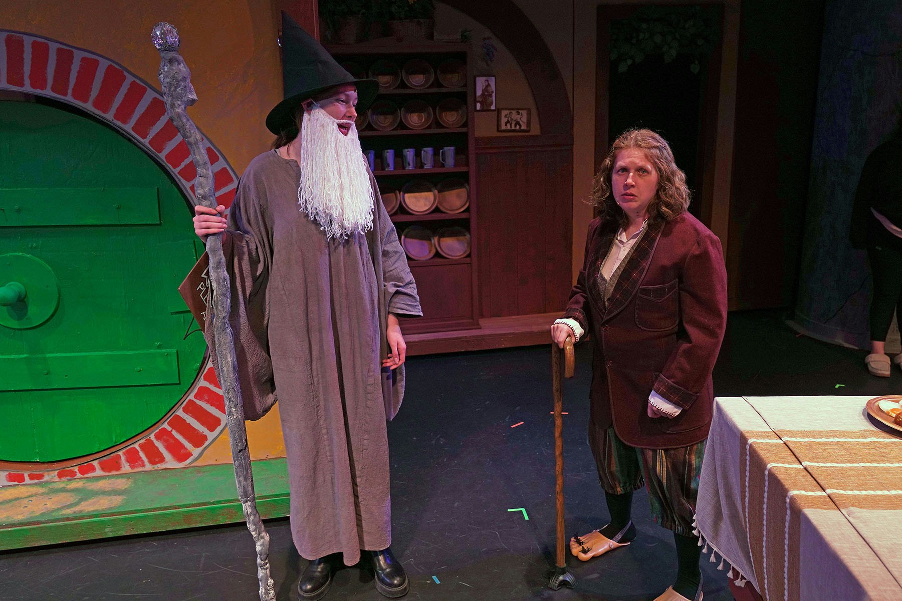 Two people in a play: one in a wizard costume with a staff, the other in a medieval outfit with a cane, standing in front of a hobbit door.