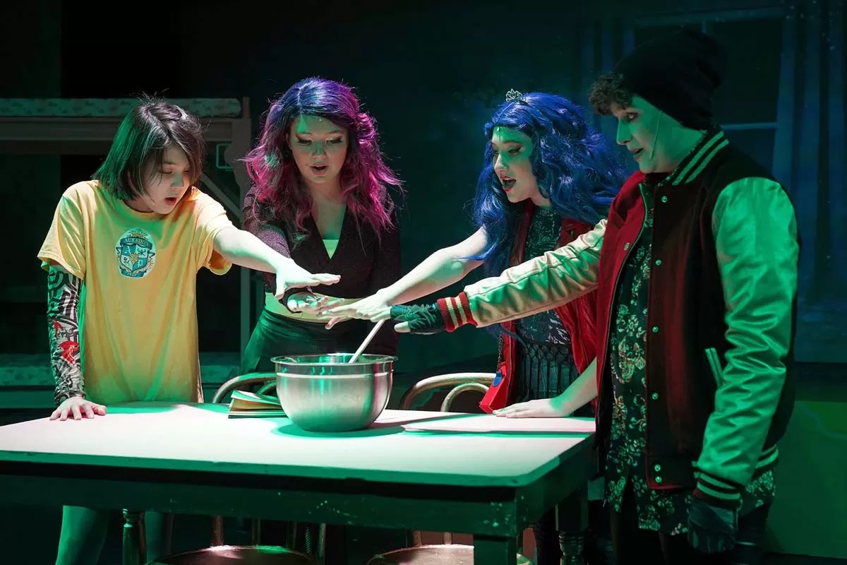 Four people with colorful hair stand around a table, hands over a bowl, as if performing a ritual.