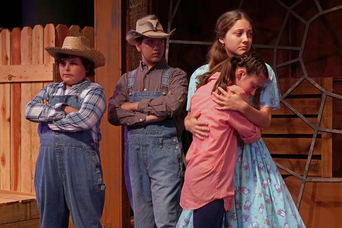 A group of kids in cowboy hats and overalls; two hug, one is sad, and two have arms crossed, in a rustic setting.