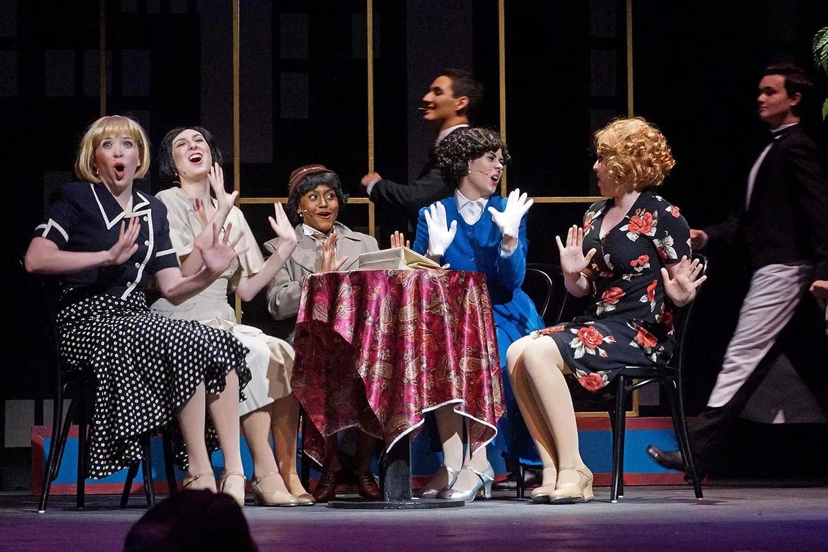 Five people at a table on stage, dressed in vintage clothing, expressing surprise or excitement.