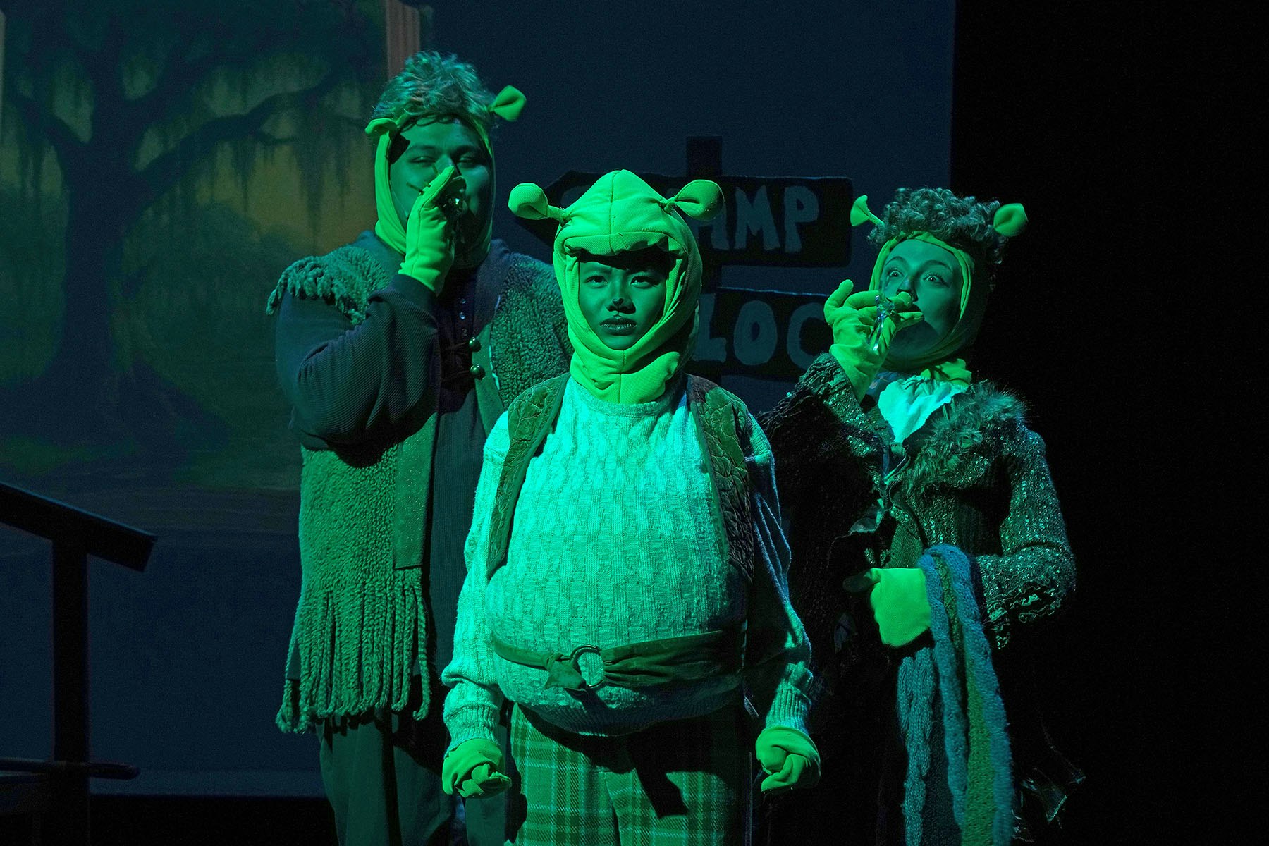 Three people in green costumes with ogre-like ears on stage, one holding a flute.