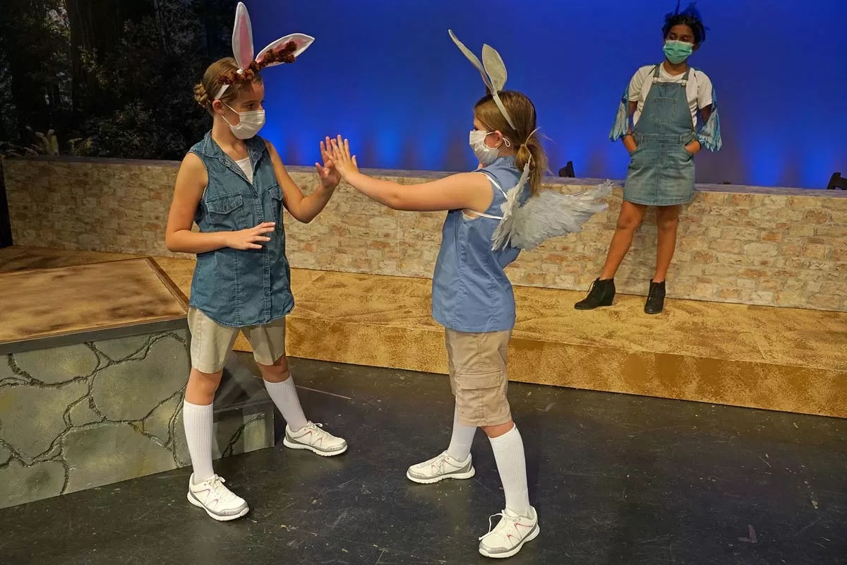 Children in costume with masks, one wearing bunny ears and another with wings, are performing on stage with a blue background.