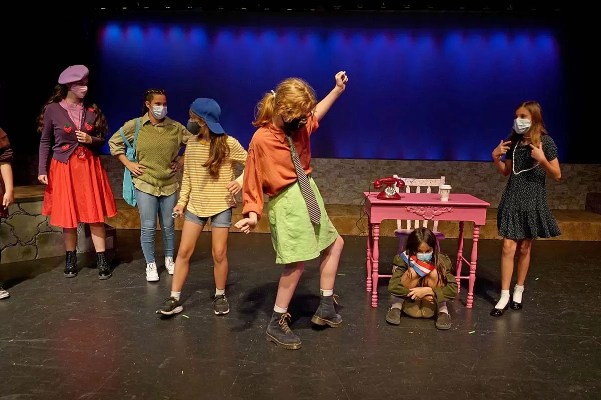 Children in a play wearing costumes and masks, acting on stage with a colorful backdrop, table, and chairs.