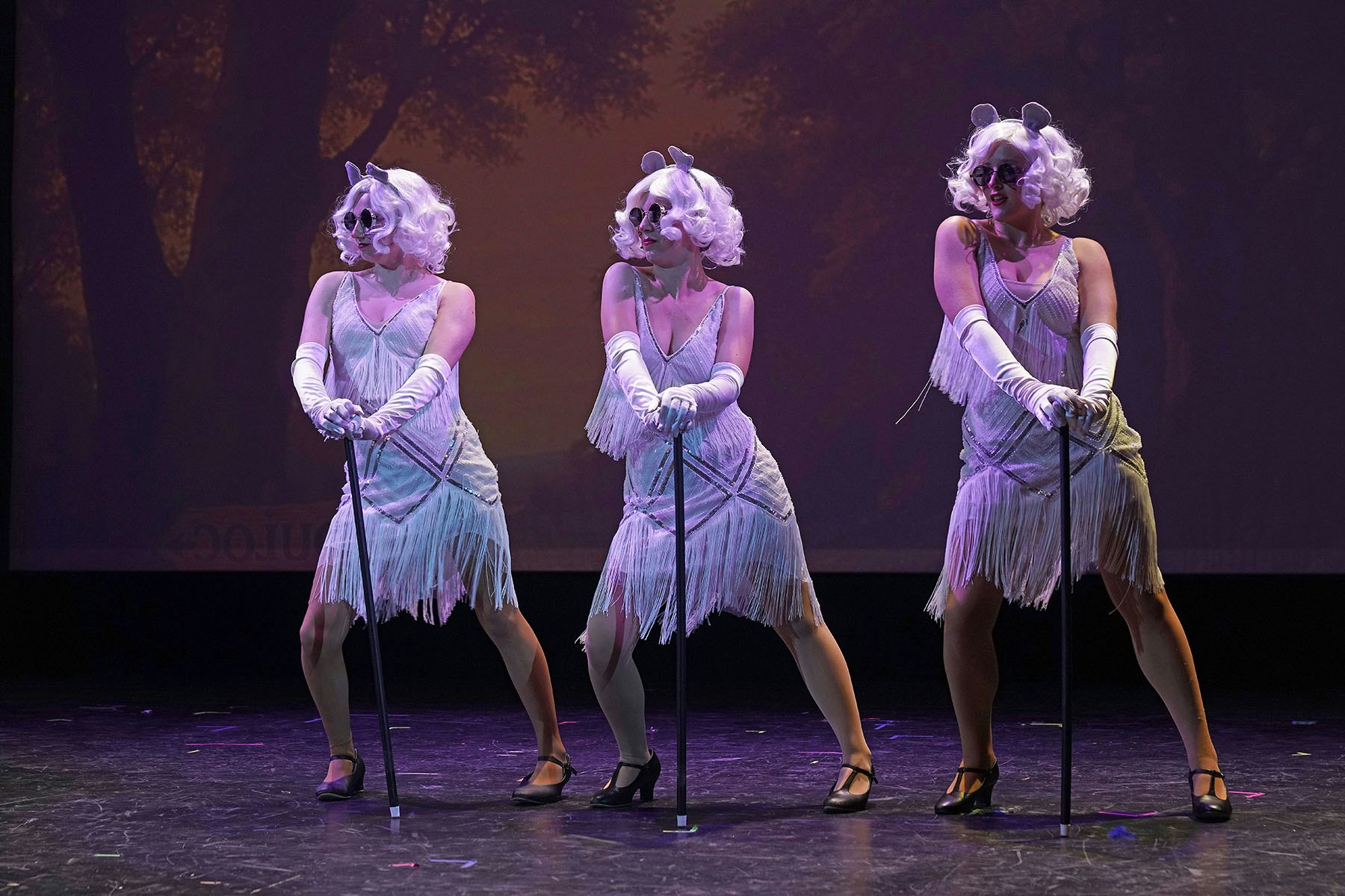 Three performers in fringe dresses, gloves, sunglasses, and wigs on stage holding canes, performing in sync.