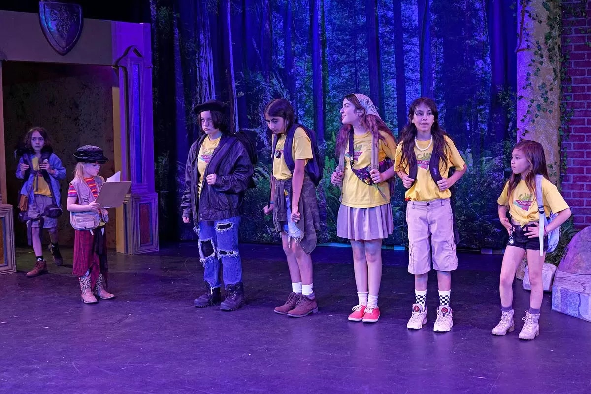 Children on stage in a play, wearing yellow shirts and backpacks, with a forest backdrop.
