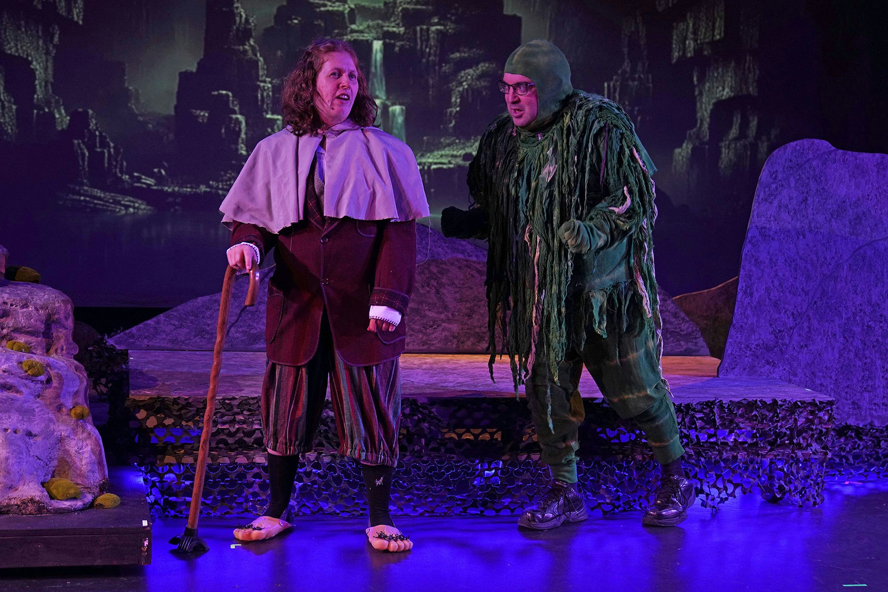 Two actors perform on stage, one in period clothing with a cane, the other in a green, moss-like costume.