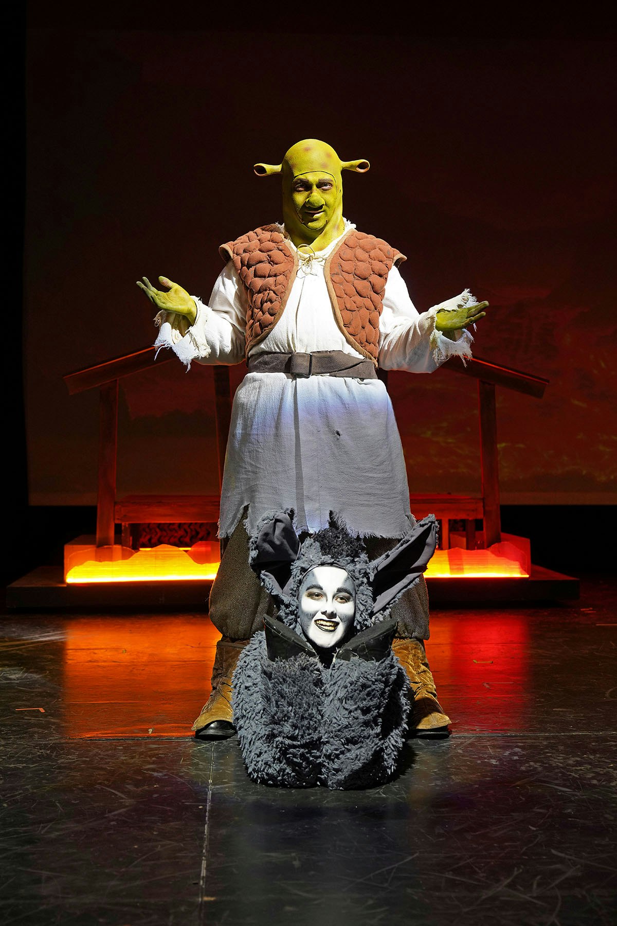 Two people in costume on stage; one as a green ogre, the other a gray creature with big ears, resembling a theatrical production.