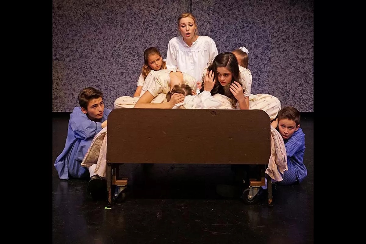 A group of children in pajamas huddle around a bed on stage, with a woman in white singing or speaking.