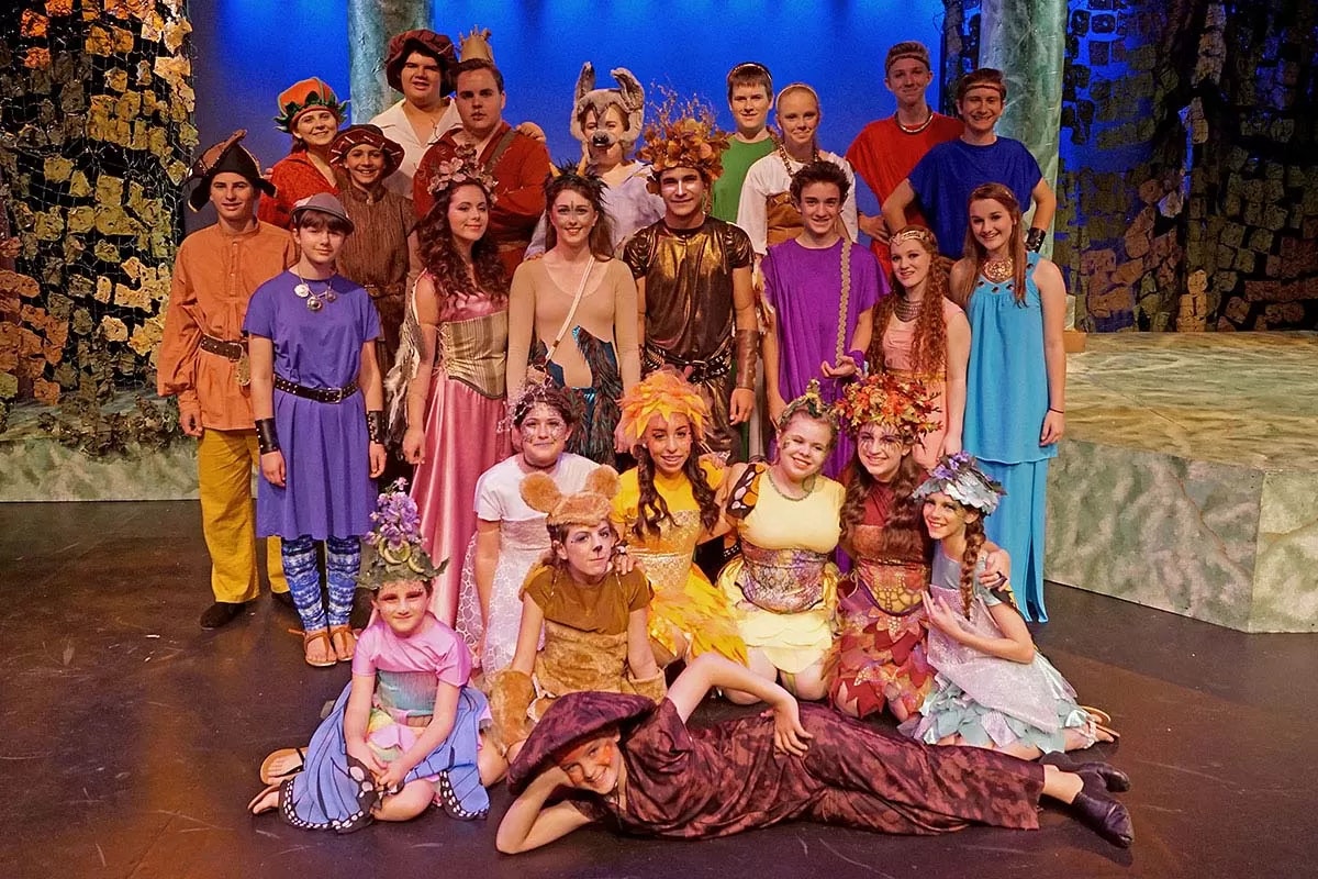 A group of people in colorful, theatrical costumes pose on stage, suggesting a play or performance setting.