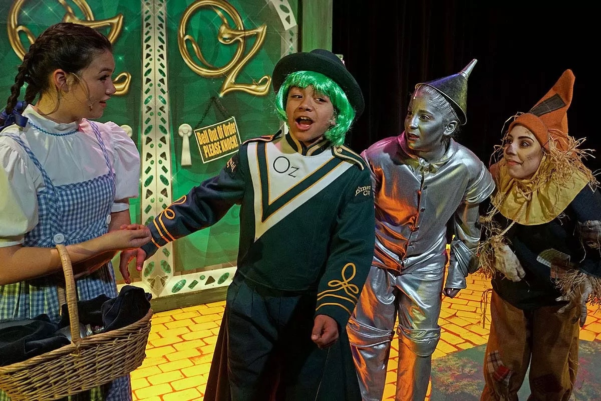 Four people in Oz-themed costumes on a stage: Dorothy, a green-haired character, Tin Woodman, and Scarecrow near an "Oz" sign.