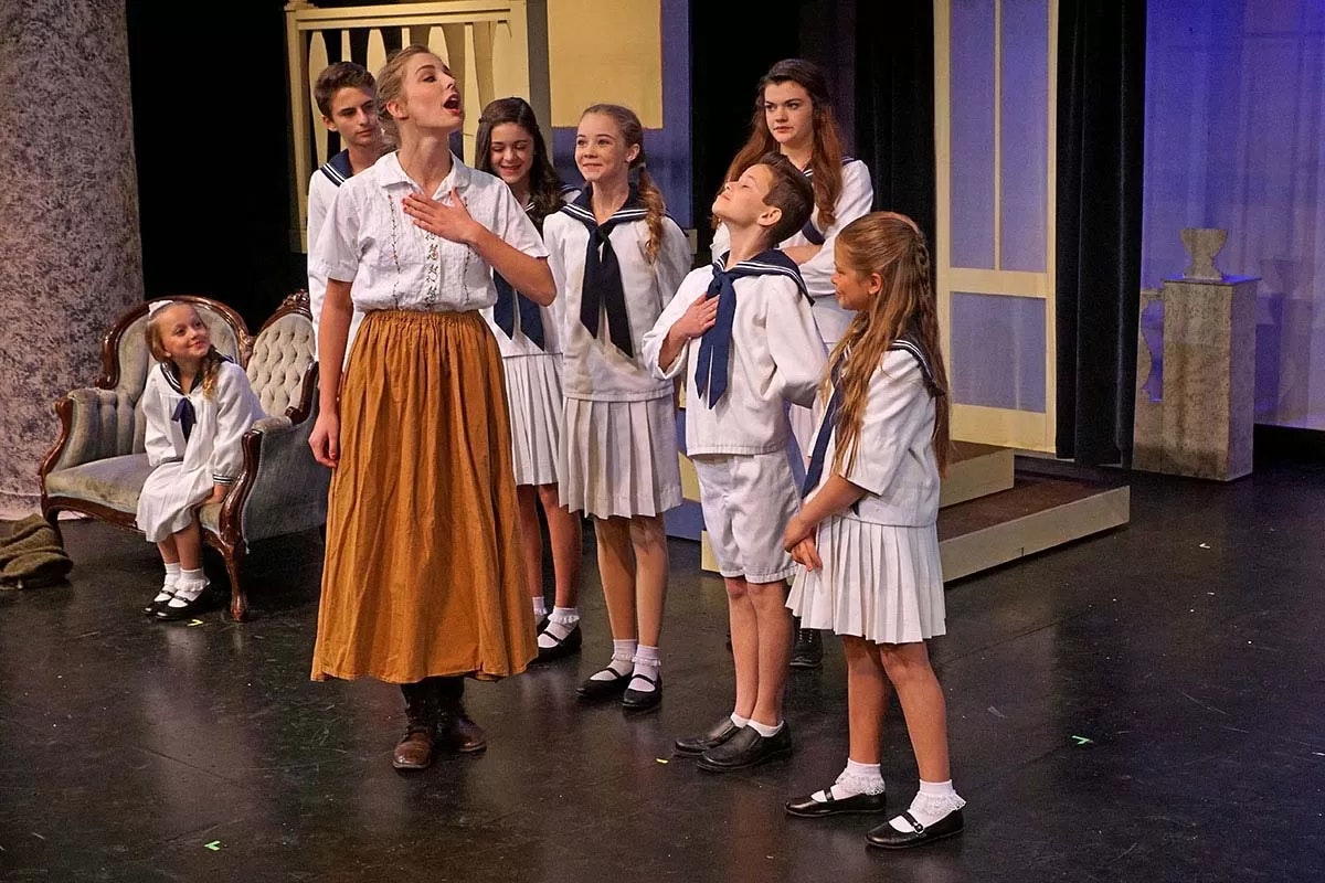 A woman and children on stage in sailor-style outfits, possibly performing a scene from a play or musical.